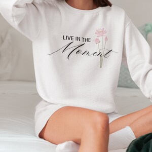 May include: White sweatshirt with the text "LIVE IN THE Moment" in black script and a pink floral design. The sweatshirt is a crew neck style.