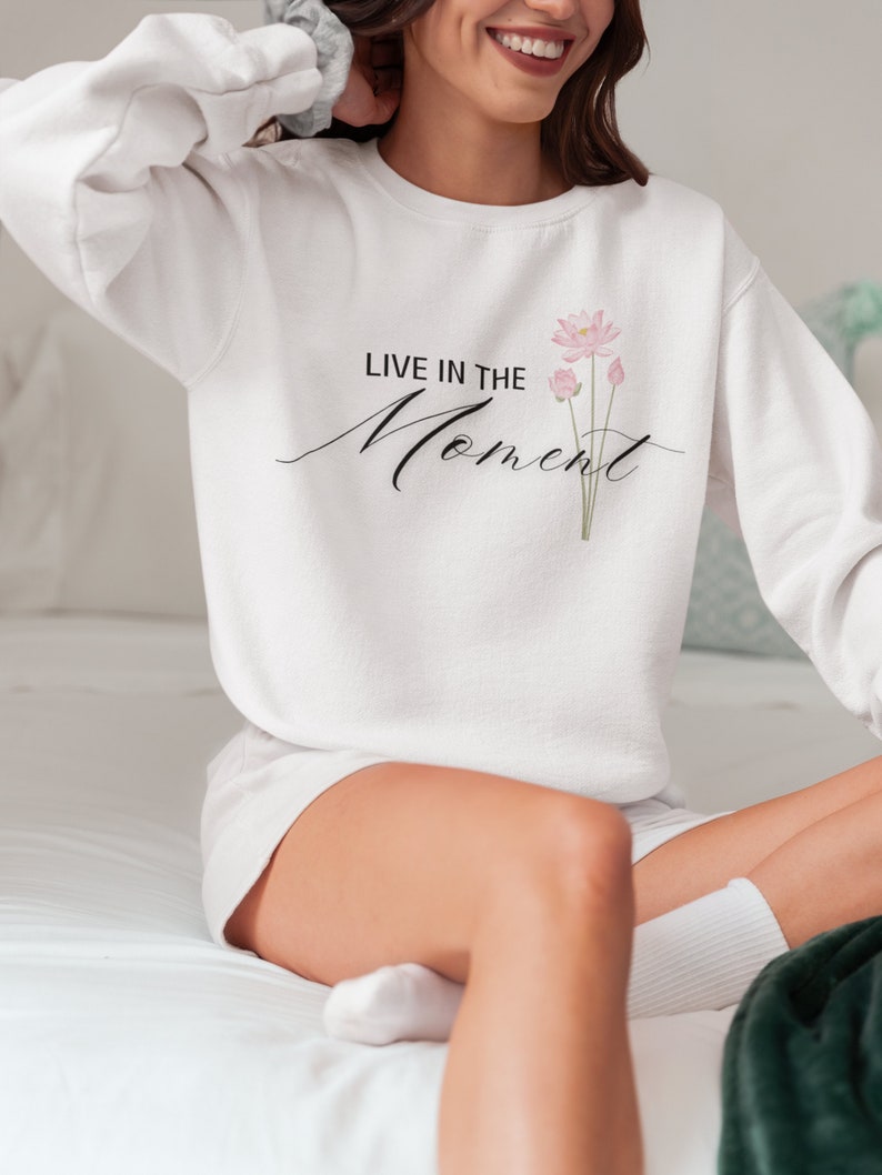 May include: White sweatshirt with the text "LIVE IN THE Moment" in black script and a pink floral design. The sweatshirt is a crew neck style.