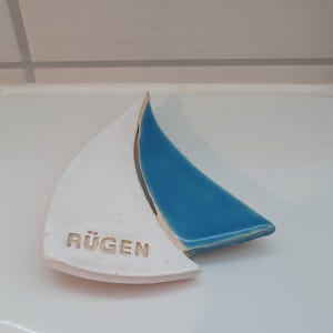 Könnte beinhalten: Ein keramisches Deko-Objekt in Form eines Segelbootes. Ein Segel ist weiß, das andere blau. Das Wort "RÜGEN" ist auf dem weißen Segel geprägt. Das Objekt befindet sich auf einer weißen Oberfläche.