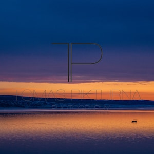 May include: A lone boat sits on a still lake at sunset. The sky is a gradient of blue, purple, and orange. The water reflects the colors of the sky. The text "TOMAS PIKTURNA PHOTOGRAPHY" is visible in the lower center of the image.