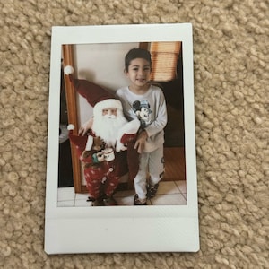 May include: A young child wearing a Mickey Mouse shirt stands next to a Santa Claus figure. The Santa Claus figure is holding a teddy bear and a cup.