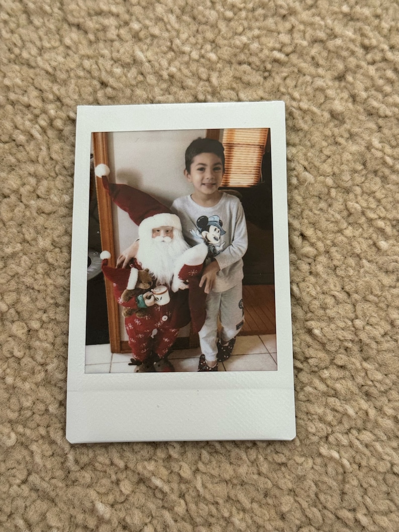 May include: A young child wearing a Mickey Mouse shirt stands next to a Santa Claus figure. The Santa Claus figure is holding a teddy bear and a cup.