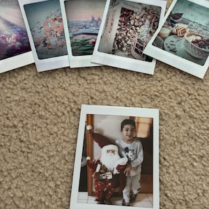 May include: A Polaroid photo of a young person standing next to a Santa Claus figure. The person is wearing a light blue shirt with a Mickey Mouse design and white pants. The Santa Claus figure is wearing a red and white suit with a white beard.