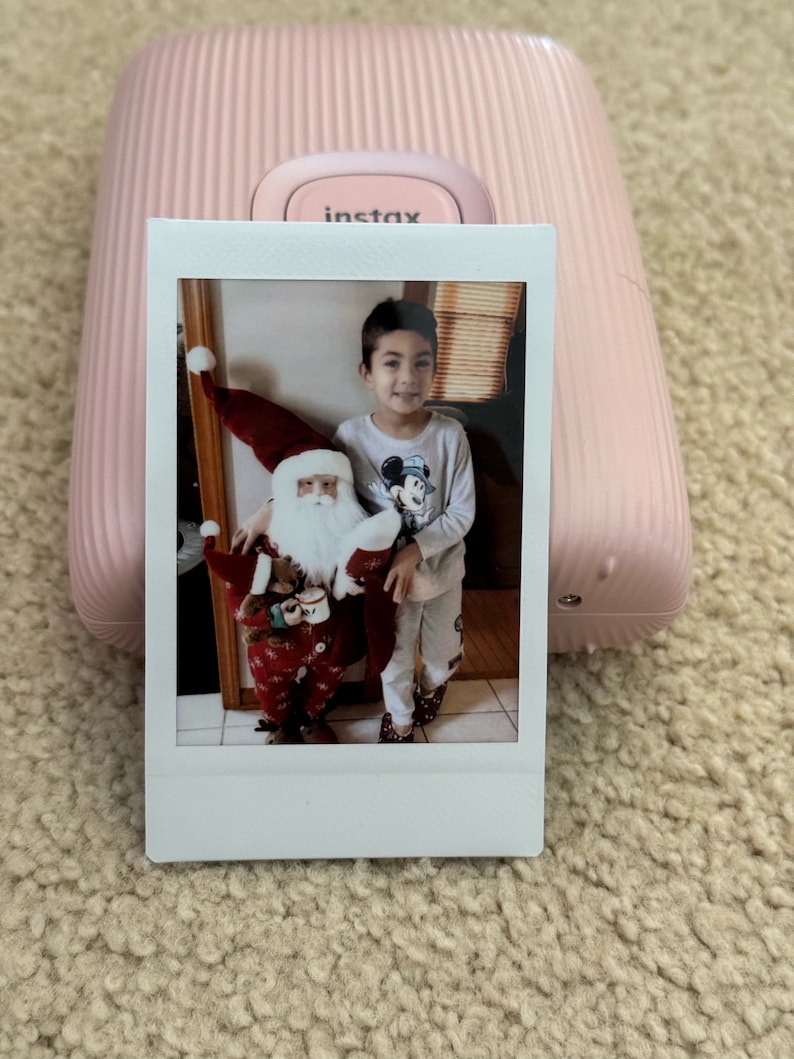 May include: A white instant camera with a pink case and the word "instax" printed on the front. A printed photo shows a young boy in a grey shirt and red pants standing next to a Santa Claus doll.