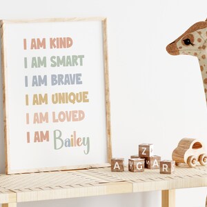 K&ouml;nnte beinhalten: Ein gerahmter Druck mit dem Text "Ich bin freundlich, ich bin klug, ich bin mutig, ich bin einzigartig, ich bin geliebt, ich bin Bailey" in bunten Buchstaben auf wei&szlig;em Hintergrund. Der Druck steht auf einem Holzregal mit einer Stoffgiraffe und Holzkl&ouml;tzen.