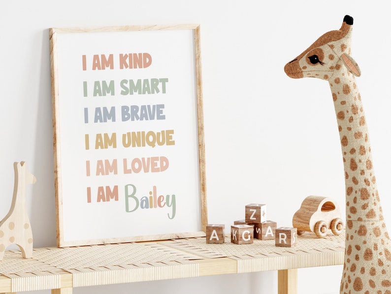 K&ouml;nnte beinhalten: Ein gerahmter Druck mit dem Text "Ich bin freundlich, ich bin klug, ich bin mutig, ich bin einzigartig, ich bin geliebt, ich bin Bailey" in bunten Buchstaben auf wei&szlig;em Hintergrund. Der Druck steht auf einem Holzregal mit einer Stoffgiraffe und Holzkl&ouml;tzen.