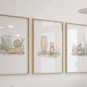 May include: Three framed watercolor prints with light wood frames. The prints feature whimsical illustrations of ocean life, including whales, surfboards, a teddy bear, and other sea creatures. The artwork has a soft, pastel color palette.