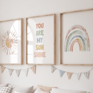 May include: Three framed watercolor prints with a sun, rainbow, and the text "You are my sunshine". The prints are hung on a white wall with a string of flags hanging below them.