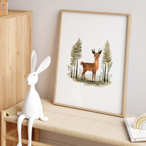 May include: A watercolor print of a deer standing in a forest with green trees. The print is framed in a light wood frame and is hanging on a white wall.