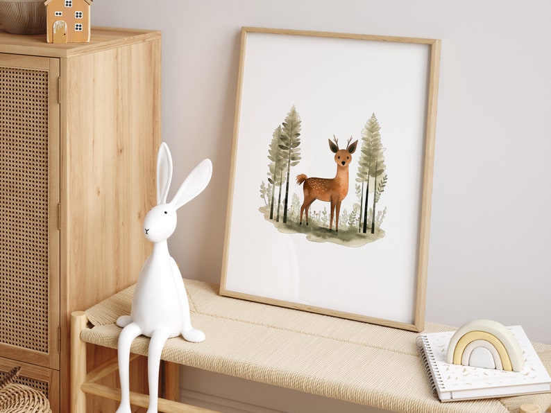 May include: A watercolor print of a deer standing in a forest with green trees. The print is framed in a light wood frame and is hanging on a white wall.