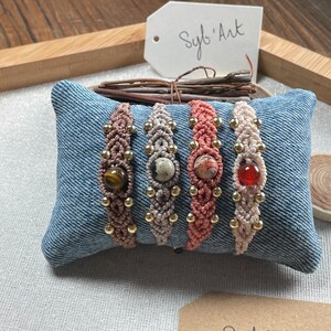 May include: Four handmade macrame bracelets with different colored cords and gemstone beads. The bracelets are displayed on a blue denim pillow. The text "Sylb' Art" is visible on a white tag.