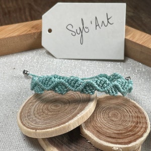 May include: A light blue macrame bracelet with silver metal beads. The bracelet is displayed on a wooden surface with a white tag that says "Syl's Art".