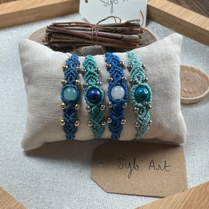 May include: Four macrame bracelets with different colored stones. The stones are aqua, lapis lazuli, white, and turquoise. The bracelets are on a white pillow with a brown tag that says "Syb' Art".