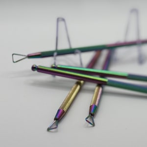 Peut inclure: Quatre outils en métal irisé avec des embouts de formes différentes, posés sur une surface blanche. Les outils sont utilisés pour le nail art et d'autres travaux manuels.
