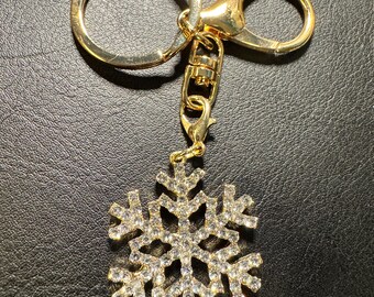 Può includere: Portachiavi in metallo dorato con un ciondolo a forma di fiocco di neve. Il ciondolo è incastonato con cristalli trasparenti e il portachiavi ha un anello rotondo e un moschettone. Accessorio a tema invernale.