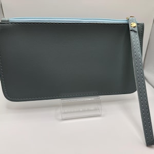 Pu&ograve; includere: Una pochette rettangolare grigio ardesia con interno azzurro e cinturino da polso abbinato. La pochette ha un tirazip dorato ed &egrave; esposta su un supporto in acrilico trasparente. Il cinturino da polso &egrave; cucito.
