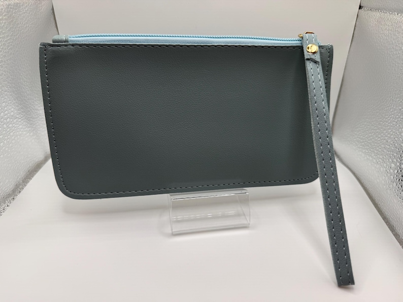 Pu&ograve; includere: Una pochette rettangolare grigio ardesia con interno azzurro e cinturino da polso abbinato. La pochette ha un tirazip dorato ed &egrave; esposta su un supporto in acrilico trasparente. Il cinturino da polso &egrave; cucito.