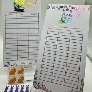 May include: Two white printable sheets with a grid pattern and a floral border. The sheets are decorated with Easter-themed stickers, including a bunny, eggs, and butterflies. The text "Happy Easter" is visible on one of the sheets.