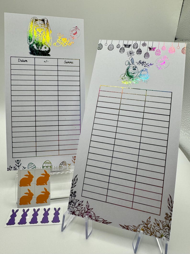 May include: Two white printable sheets with a grid pattern and a floral border. The sheets are decorated with Easter-themed stickers, including a bunny, eggs, and butterflies. The text "Happy Easter" is visible on one of the sheets.