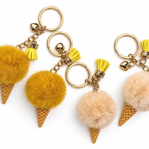 May include: Four ice cream cone keychains featuring fluffy pom-poms in mustard yellow and beige. Each keychain includes a gold-tone metal ring, a small heart charm, and a bright yellow tassel attached to a textured, waffle-patterned cone base.