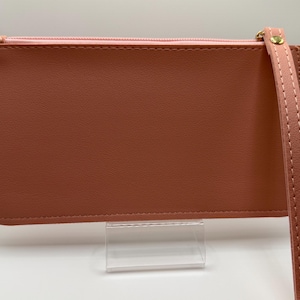 Pu&ograve; includere: Un portafoglio rettangolare color terracotta con cerniera rosa e cinturino da polso. Il portafoglio ha una consistenza liscia e un tirazip dorato. Il cinturino da polso &egrave; dello stesso colore del portafoglio ed &egrave; cucito.