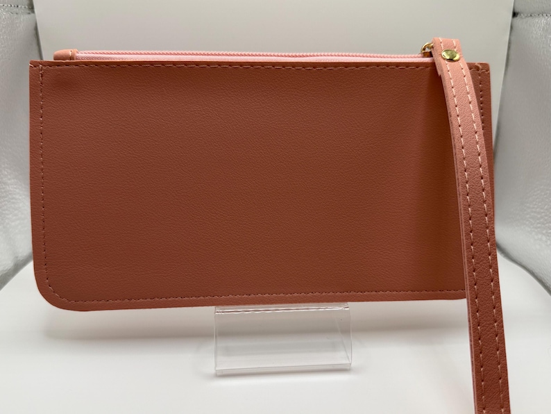 Pu&ograve; includere: Un portafoglio rettangolare color terracotta con cerniera rosa e cinturino da polso. Il portafoglio ha una consistenza liscia e un tirazip dorato. Il cinturino da polso &egrave; dello stesso colore del portafoglio ed &egrave; cucito.