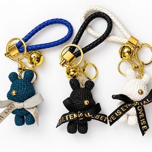 May include: Three keychains featuring crocheted rabbit figures in dark blue, black, and white. Each rabbit has a gold bead eye and a ribbon tied around its neck with the text LOVE IS ETERNAL. The keychains have braided loops and gold hardware.