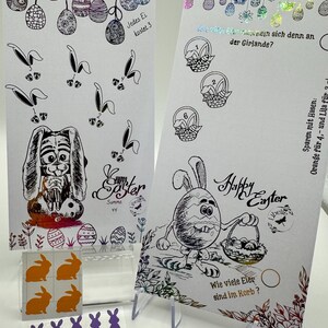 May include: Two printable Easter cards with bunny illustrations and a basket of eggs. The cards are decorated with colorful foil accents. The text on the cards reads "Happy Easter" and "Jedes Ei kostet 3". There are also stickers of purple and orange bunnies.