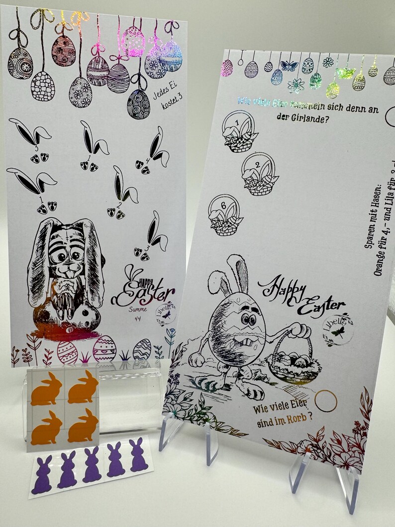 May include: Two printable Easter cards with bunny illustrations and a basket of eggs. The cards are decorated with colorful foil accents. The text on the cards reads "Happy Easter" and "Jedes Ei kostet 3". There are also stickers of purple and orange bunnies.