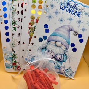 May include: A collection of winter-themed planner inserts and accessories. The inserts feature festive designs with snowflakes, gnomes, and the text "hello Winter." A small bag contains several red, cylindrical items.