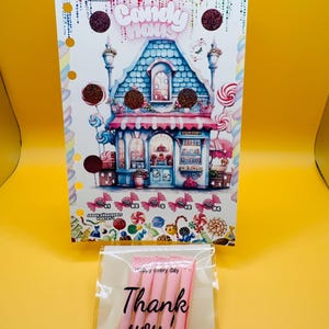 Pode incluir: Um cart&atilde;o colorido com tema de doces com uma ilustra&ccedil;&atilde;o de uma loja de doces, com um telhado azul e detalhes em rosa. O cart&atilde;o tem o t&iacute;tulo "Candy House" e &eacute; decorado com doces e glitter. Uma sacola transparente com clipes rosa e o texto "Thank you!" est&aacute; na frente.