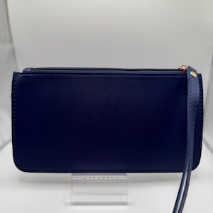 Pu&ograve; includere: Un portafoglio a pochette blu navy con cinturino da polso. Il portafoglio rettangolare ha una chiusura a cerniera con un tirante color oro. Il cinturino da polso &egrave; attaccato al lato del portafoglio. Il portafoglio &egrave; esposto su un supporto in acrilico trasparente.