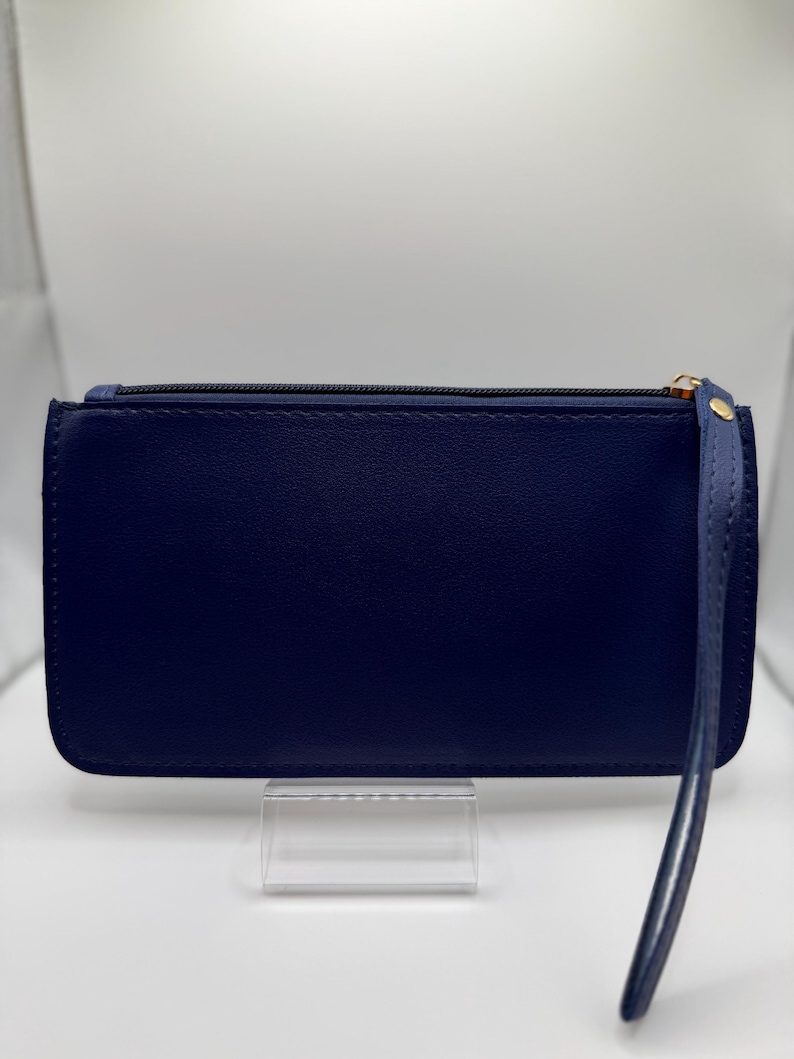Pu&ograve; includere: Un portafoglio a pochette blu navy con cinturino da polso. Il portafoglio rettangolare ha una chiusura a cerniera con un tirante color oro. Il cinturino da polso &egrave; attaccato al lato del portafoglio. Il portafoglio &egrave; esposto su un supporto in acrilico trasparente.