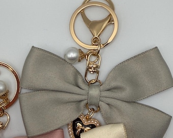 Può includere: Un portachiavi dorato con un fiocco grigio, un accento di perla e un ciondolo a forma di cuore. Il ciondolo a forma di cuore è nero con un design a fiocco dorato ed è circondato da piccole perle. Il portachiavi ha un moschettone e un anello diviso.