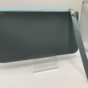 Pu&ograve; includere: Una pochette grigio ardesia con cerniera blu chiaro e cinturino da polso. La pochette ha un tirazip dorato e un cinturino da polso abbinato. Le cuciture sono visibili sui bordi della pochette e del cinturino. La pochette &egrave; esposta su un supporto in acrilico trasparente.