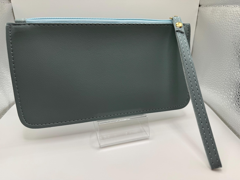 Pu&ograve; includere: Una pochette grigio ardesia con cerniera blu chiaro e cinturino da polso. La pochette ha un tirazip dorato e un cinturino da polso abbinato. Le cuciture sono visibili sui bordi della pochette e del cinturino. La pochette &egrave; esposta su un supporto in acrilico trasparente.