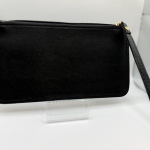 Pu&ograve; includere: Una pochette rettangolare nera con un cinturino da polso. La pochette ha una chiusura a cerniera con un tirante color oro. Il cinturino da polso &egrave; attaccato al lato della pochette. La pochette &egrave; esposta su un supporto in acrilico trasparente.