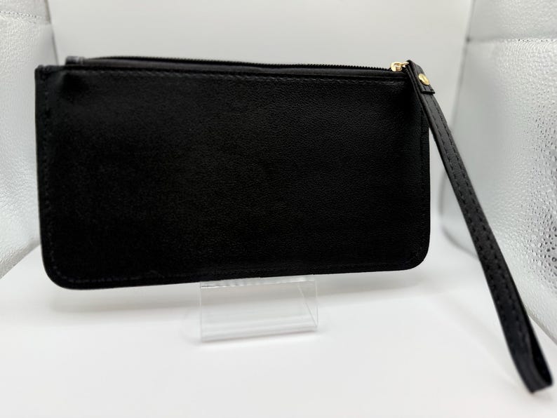 Pu&ograve; includere: Una pochette rettangolare nera con un cinturino da polso. La pochette ha una chiusura a cerniera con un tirante color oro. Il cinturino da polso &egrave; attaccato al lato della pochette. La pochette &egrave; esposta su un supporto in acrilico trasparente.