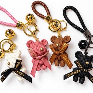 May include: Four bear-shaped keychains with braided wrist straps in white, pink, brown, and black. Each bear features a textured surface, a gold or black bell, and a ribbon tied around the neck with the text LOVE ETERNAL printed in gold letters.