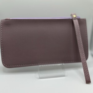 Pu&ograve; includere: Una pochette color malva con cinturino da polso. Il portafoglio rettangolare ha una chiusura a cerniera e un interno lilla chiaro. Il cinturino da polso &egrave; fissato con un rivetto color oro. La pochette &egrave; esposta su un supporto trasparente.