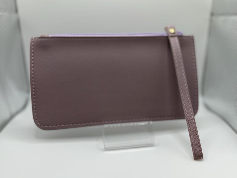 Pu&ograve; includere: Una pochette color malva con cinturino da polso. Il portafoglio rettangolare ha una chiusura a cerniera e un interno lilla chiaro. Il cinturino da polso &egrave; fissato con un rivetto color oro. La pochette &egrave; esposta su un supporto trasparente.