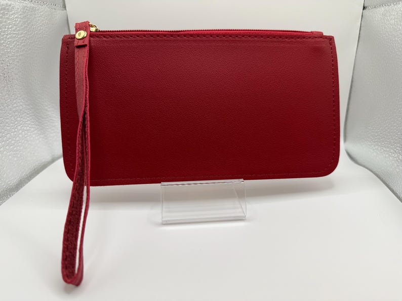 Pu&ograve; includere: Un portafoglio in pelle rosso con cinturino da polso. Il portafoglio rettangolare ha una chiusura con cerniera e un tirante dorato. Il cinturino da polso &egrave; attaccato al lato del portafoglio. Il portafoglio &egrave; esposto su un supporto in acrilico trasparente.