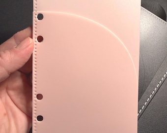 Può includere: Un inserto per agenda in plastica rosa pallido con una fila verticale di sei fori circolari lungo il bordo sinistro per gli anelli. Il foglio rettangolare presenta un design a tasca curva sulla parte anteriore con bordo cucito.
