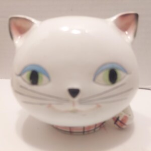 May include: A white ceramic cat figurine with blue eye shadow, green eyes, and a black nose. The cat has pink inner ears and whiskers. A pink and white plaid pattern is visible on the base and paw.
