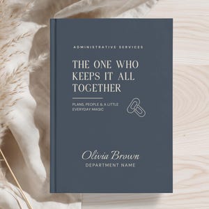 May include: A dark blue hardcover notebook with white text that reads ADMINISTRATIVE SERVICES, THE ONE WHO KEEPS IT ALL TOGETHER, PLANS, PEOPLE & A LITTLE EVERYDAY MAGIC, Olivia Brown, and DEPARTMENT NAME, featuring a small paperclip icon.