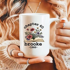 May include: White ceramic mug with the text "chapter 60 brooke -1966" and a floral design. The floral design includes pink, purple, and white flowers with green leaves. The mug has a large handle.