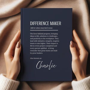 May include: A navy blue notebook with the words "DIFFERENCE MAKER" in white, along with a definition and the name "Charlie." The notebook is held by a person.
