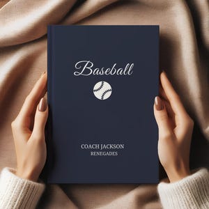 May include: A navy blue notebook with the word "Baseball" in elegant script and a baseball graphic. The text "COACH JACKSON RENEGADES" is printed below. The notebook is held by a person.