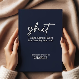 May include: A navy blue notebook with the word "shit" in white script. The text below reads "I Think About at Work But Can't Say Out Loud" and "Office Confessions CHARLIE". The notebook is held by a person wearing a cream-colored sweater.