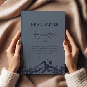 May include: A blue journal with the words "NEW CHAPTER" and "Brandon OFFICIALLY RETIRED 2025" printed on the cover. The journal has a mountain and tree design at the bottom. The journal is held by a person.